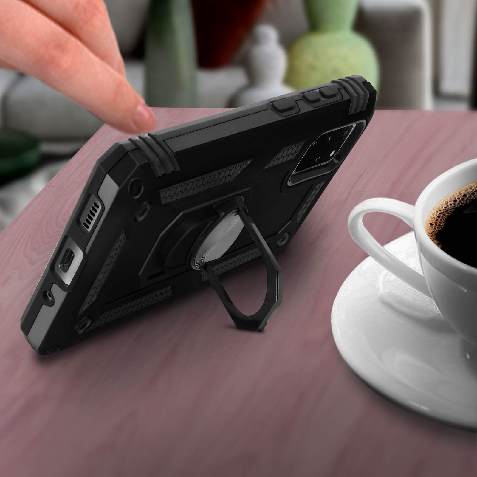 Een zwarte telefoonhoes met een ringhouder ligt op een tafel naast een kop koffie.