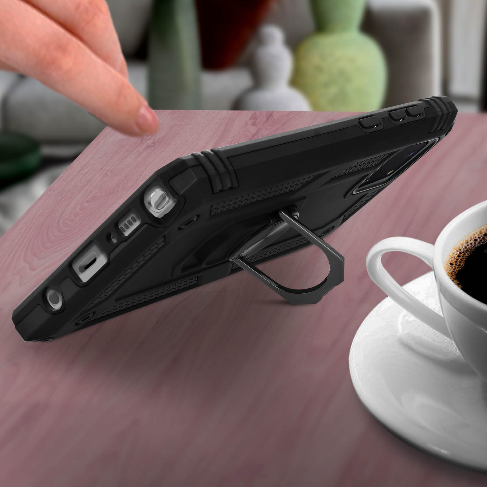 Custodia nera per telefono con supporto ad anello su un tavolo vicino a una tazza di caffè. Una mano tocca la custodia.
