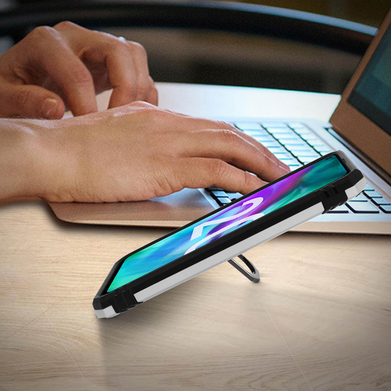 Telefono in una custodia, su una scrivania. Il telefono è appoggiato con un supporto. Una persona sta scrivendo su un laptop.