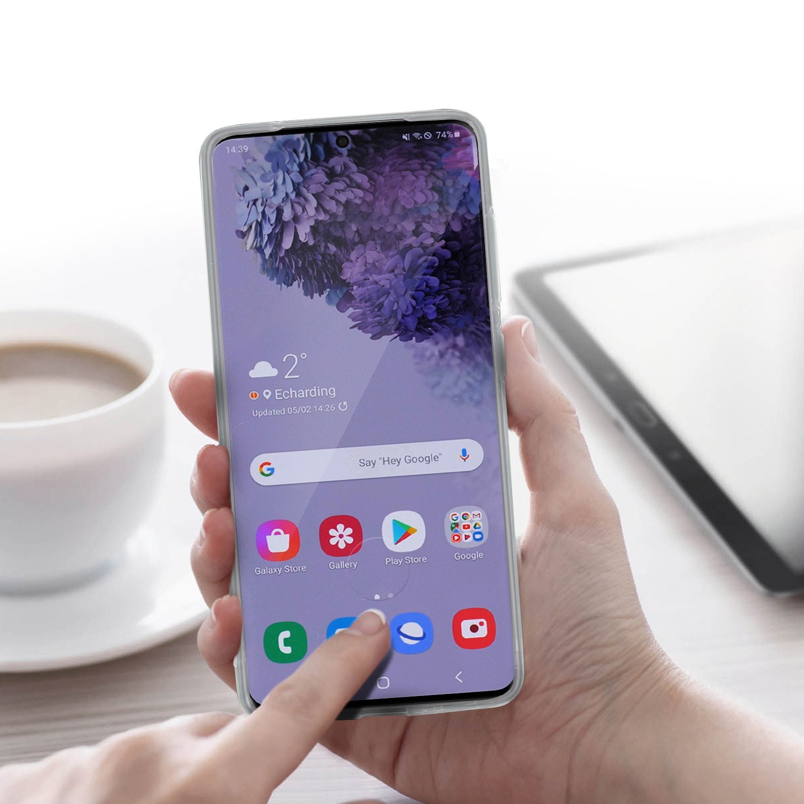 Una mano che tiene un telefono che mostra la schermata dell'app, con una tazza di caffè e un tablet sullo sfondo.