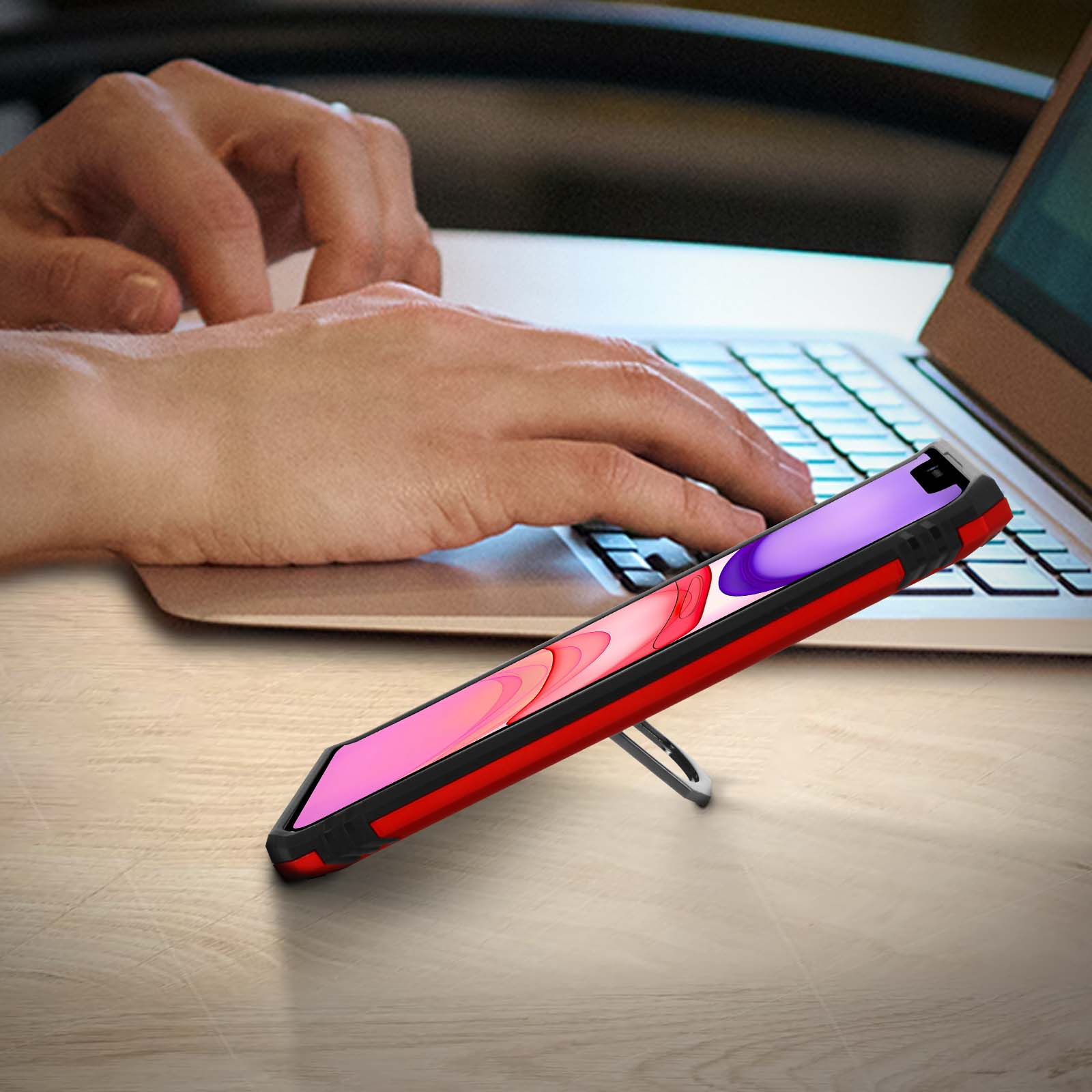 Mani che digitano su un laptop vicino a un telefono in una custodia rossa, in piedi con un supporto argentato.