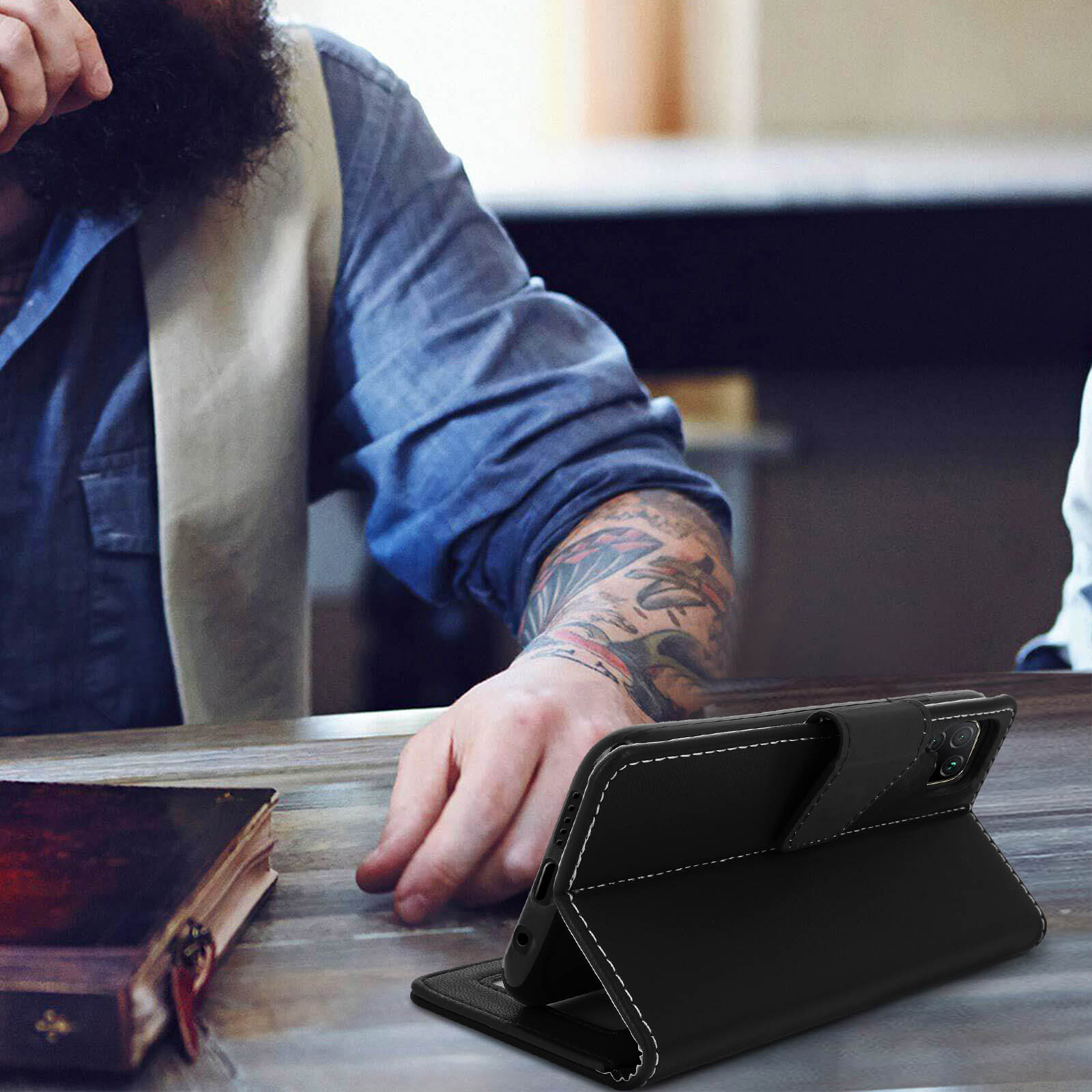 Custodia per telefono in pelle nera, aperta, con telefono all'interno. Uomo con tatuaggi sullo sfondo.