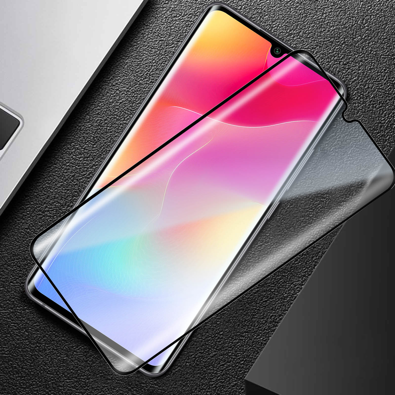 Viene mostrata una protezione per lo schermo dello smartphone, con un display a gradiente colorato. Si trova su una superficie nera strutturata, vicino a un laptop.