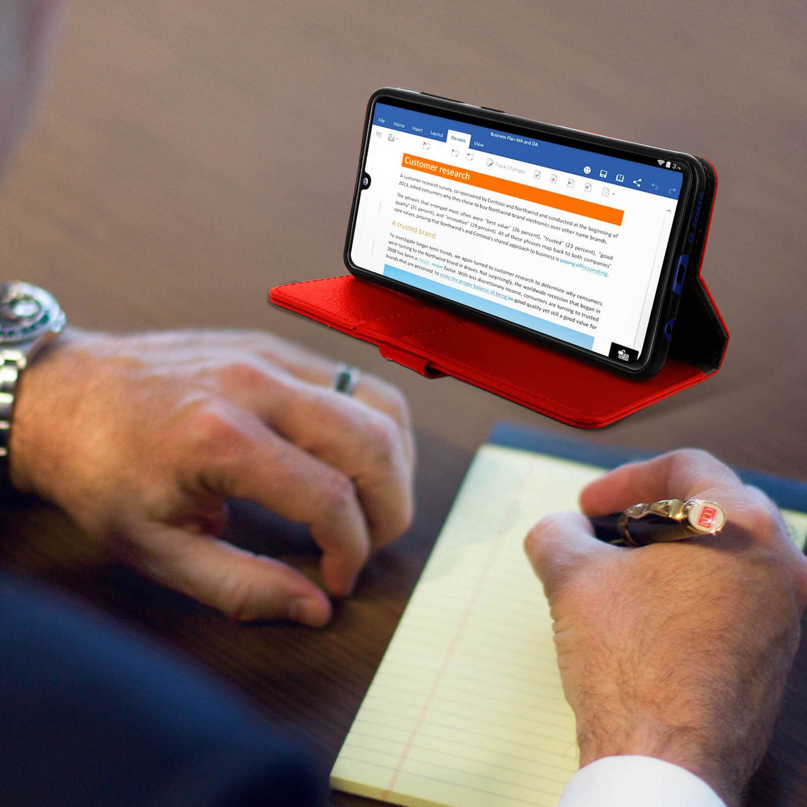 Mains tenant un téléphone dans un étui rouge, avec un document sur l'écran et un stylo écrivant sur un bloc-notes.