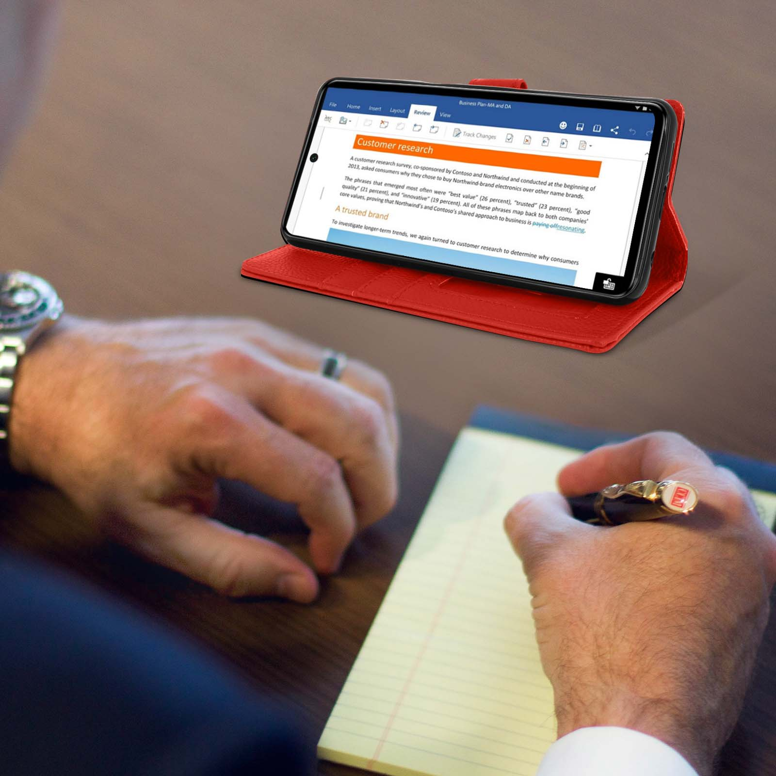 Telefoon in rode hoes op tafel. Document geopend. Hand schrijft op notitieblok. Horloge om pols.