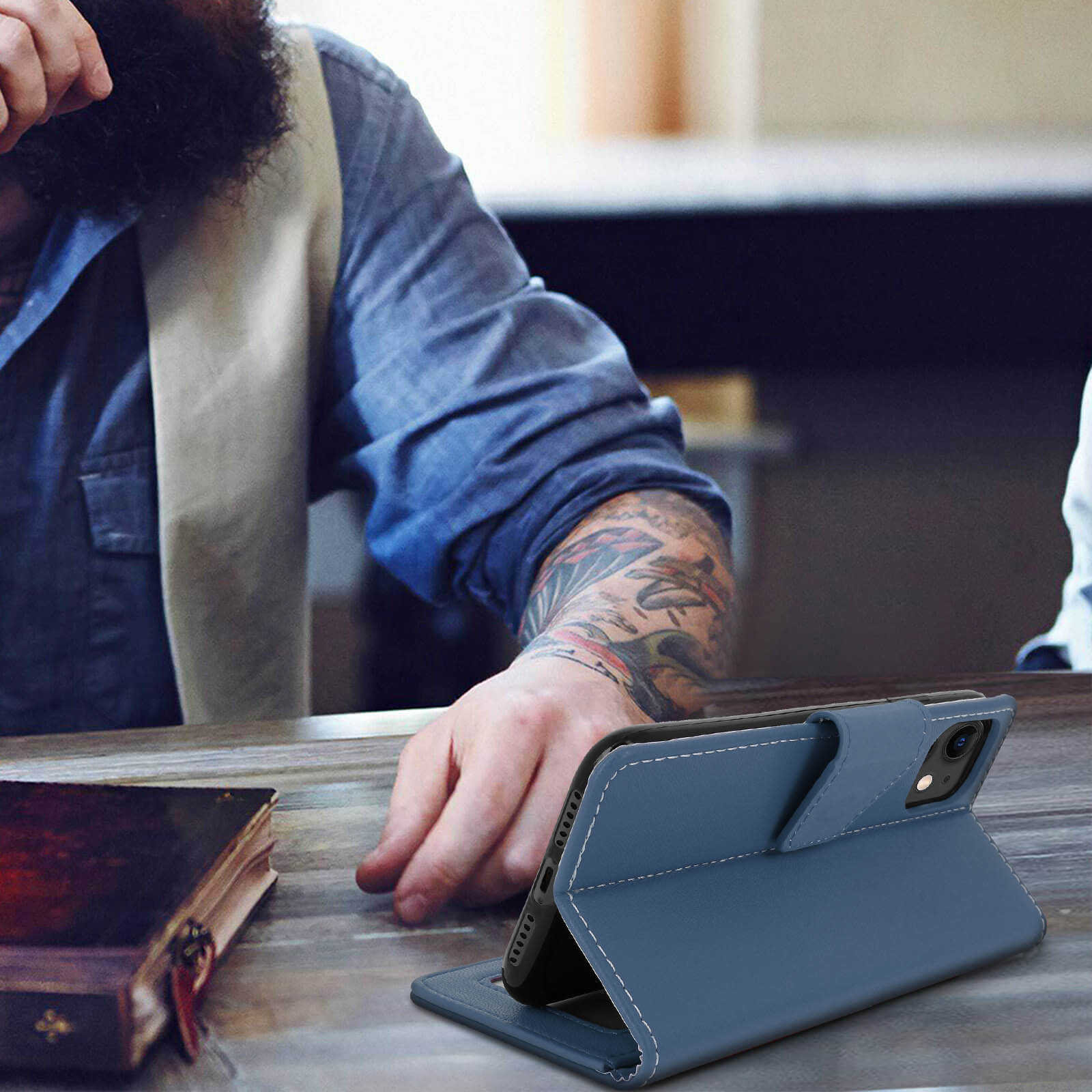 La mano di un uomo poggia su un tavolo vicino a una custodia per telefono, con un libro sullo sfondo.