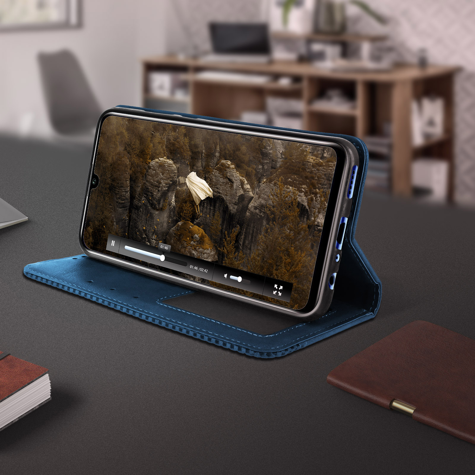 Telefono con video in custodia blu. Scrivania scura con quaderno marrone e ufficio sfocato.