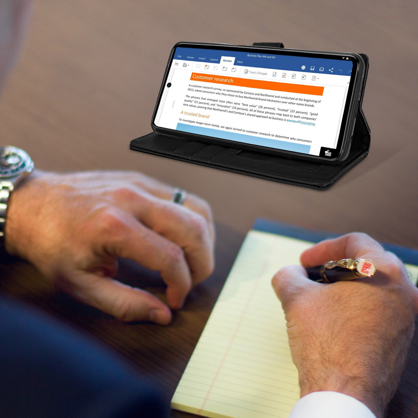 Een persoon schrijft op een notitieblok, met een telefoon die een document in een hoes weergeeft.