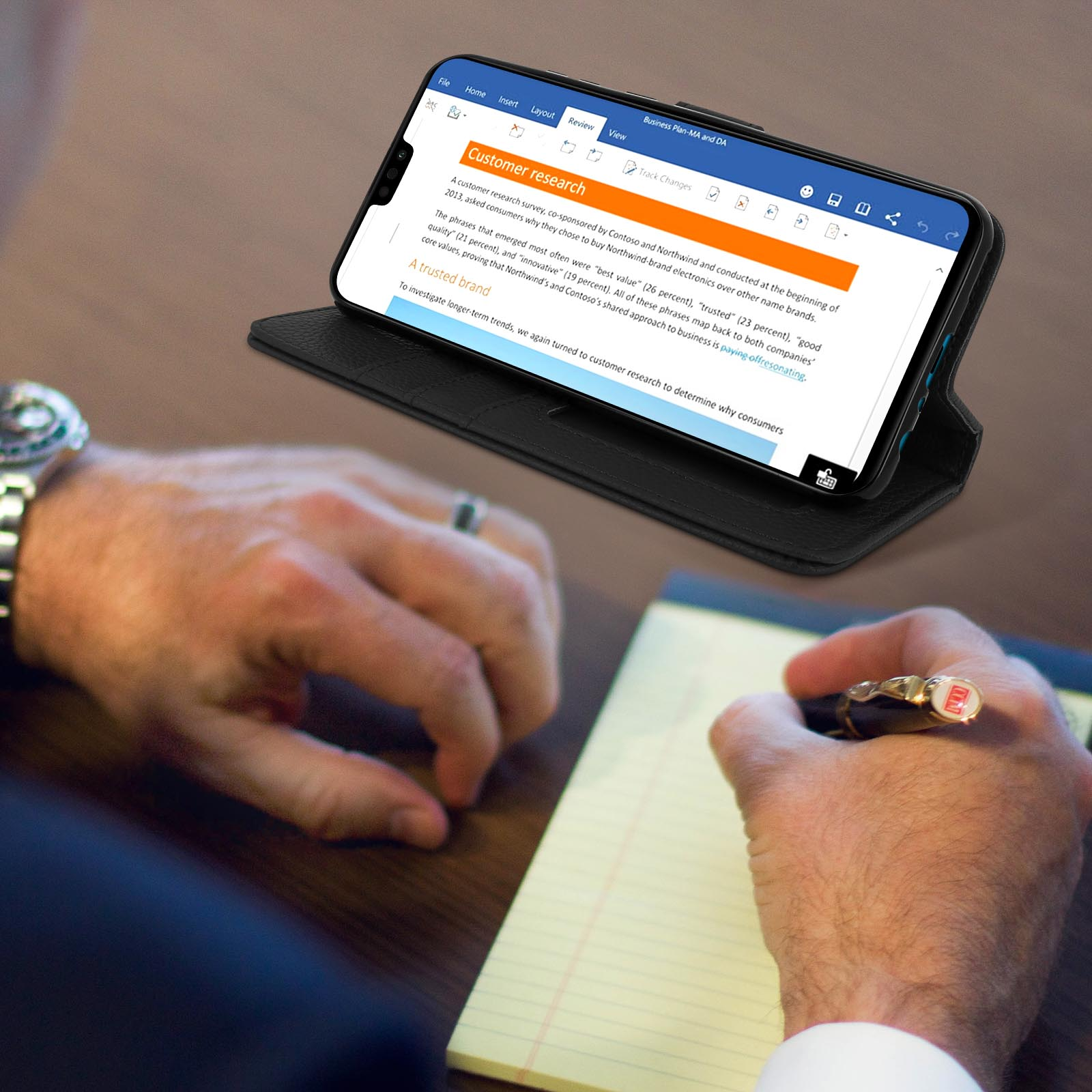 Persoon schrijft op notitieblok met telefoon in een open hoes. De telefoon toont klantenonderzoek.