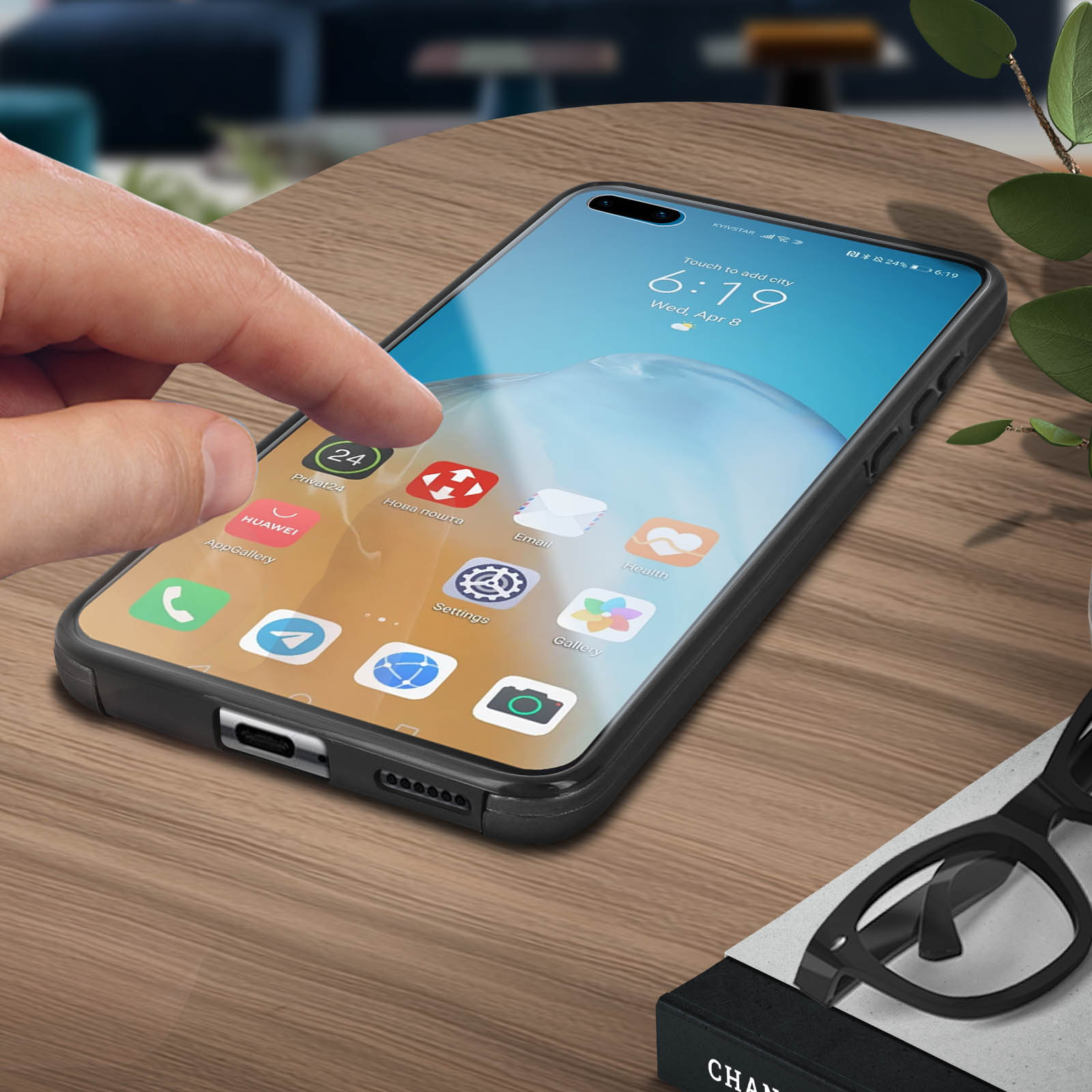 Mano tocca schermo telefono. Il telefono mostra icone. Sfondo: tavolo legno, occhiali e pianta.