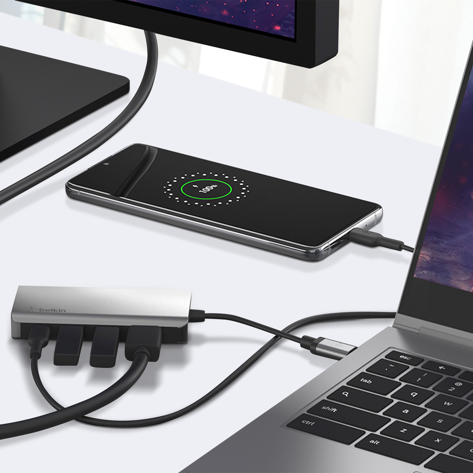 Laptop, telefono in carica e un hub Belkin su una scrivania bianca. Cavi neri visibili.