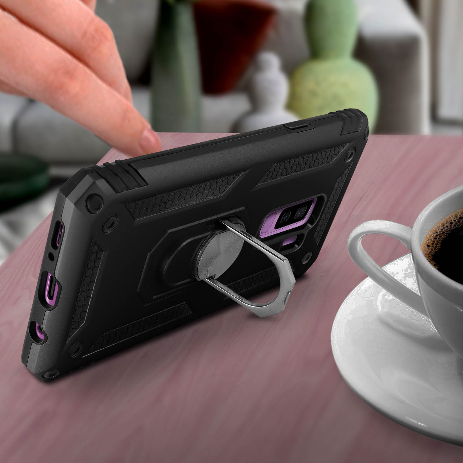 Custodia nera con supporto ad anello su tavolo rosa. Una tazza di caffè è vicina.