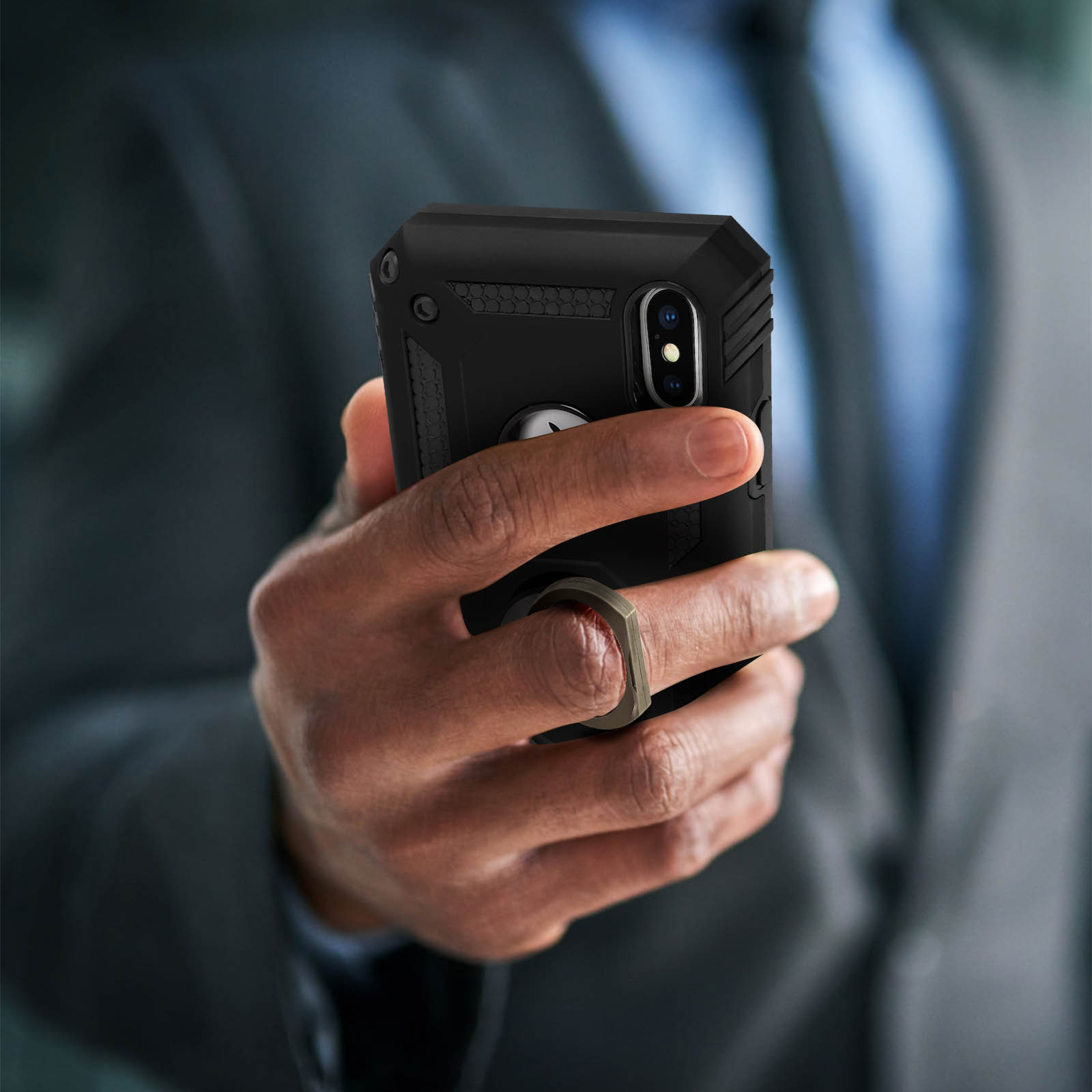 Una mano che tiene un telefono con custodia nera e supporto ad anello. Un completo è sullo sfondo.