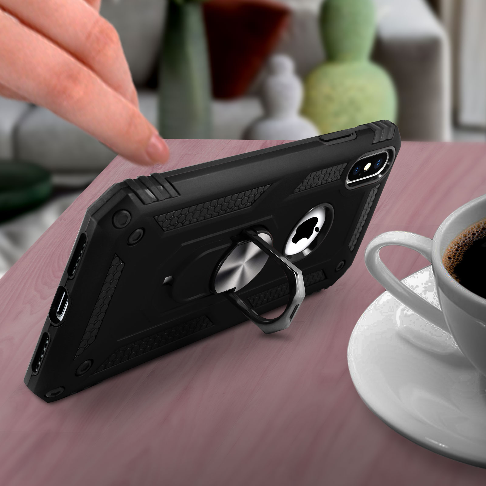 Custodia nera per telefono con supporto ad anello. Una mano raggiunge il telefono appoggiato su un tavolo con una tazza di caffè.