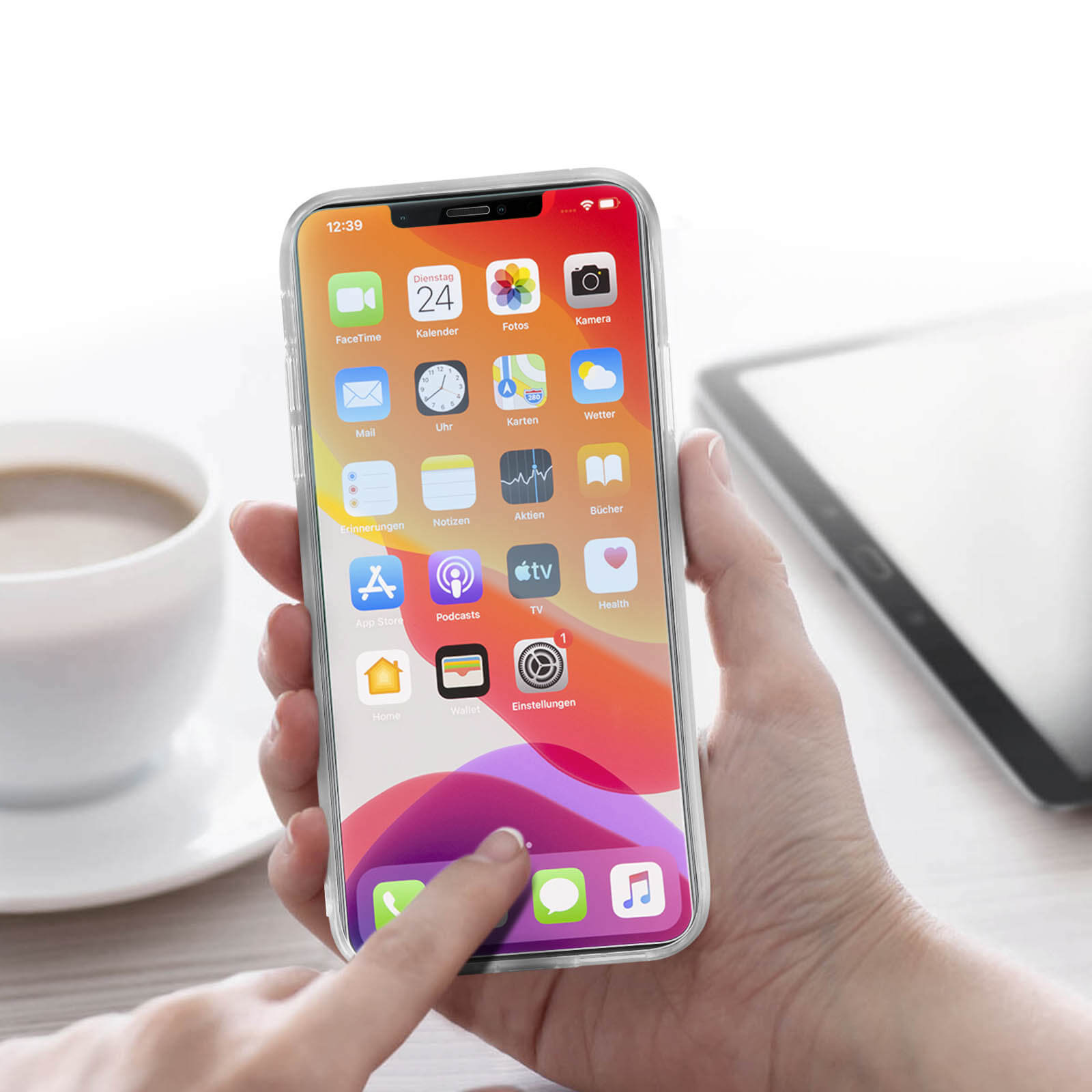 Una mano che tiene un telefono che mostra la schermata iniziale con icone di app, una tazza e un tablet sullo sfondo.