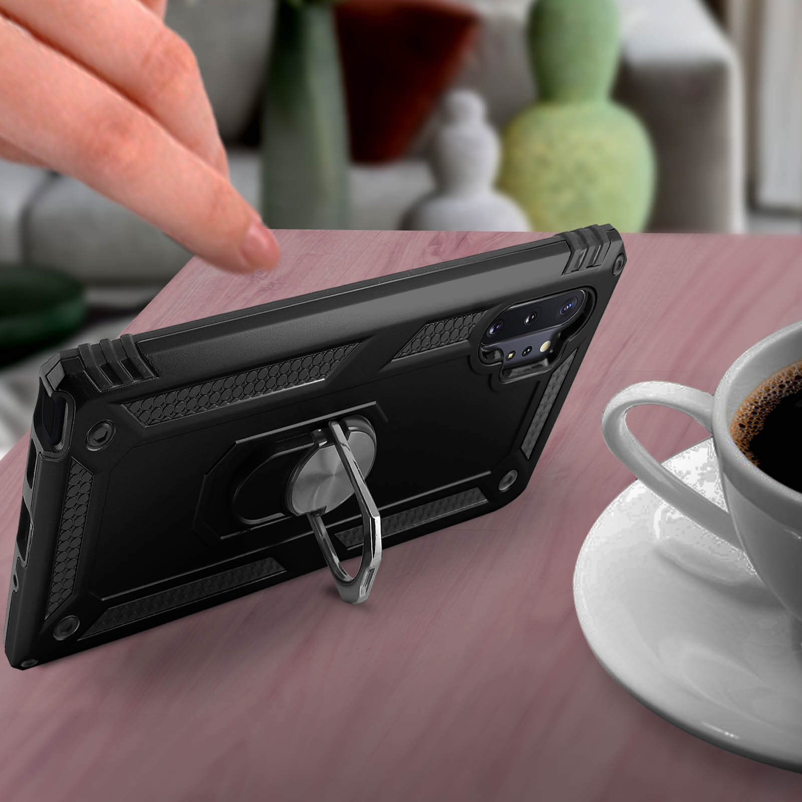 Een zwart telefoonhoesje met ringhouder op een houten tafel, met een witte kop koffie.