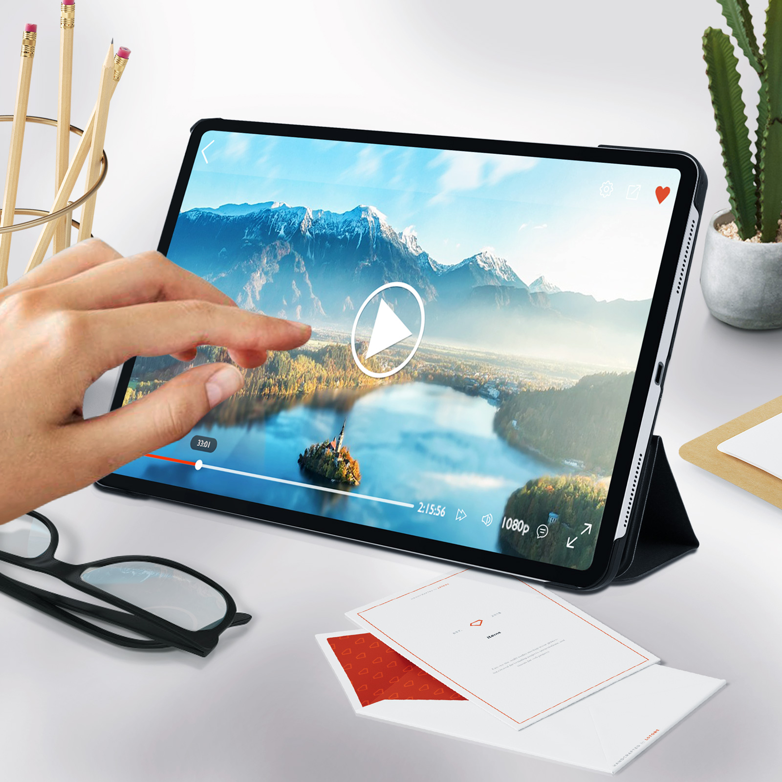 Eine Hand, die ein Tablet berührt und ein Video abspielt. Einige Gläser und Papiere liegen auf dem Tisch.