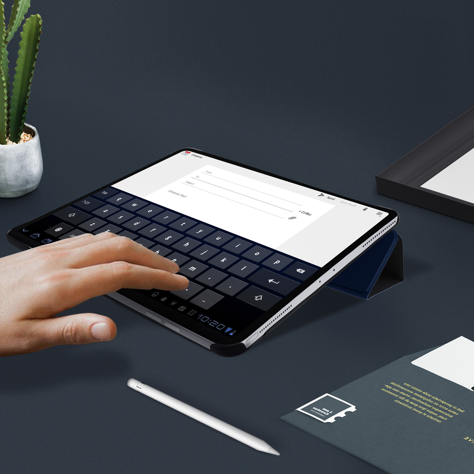 Mano che digita sulla tastiera di un tablet. Una penna bianca e un cactus sono anche sulla superficie scura.
