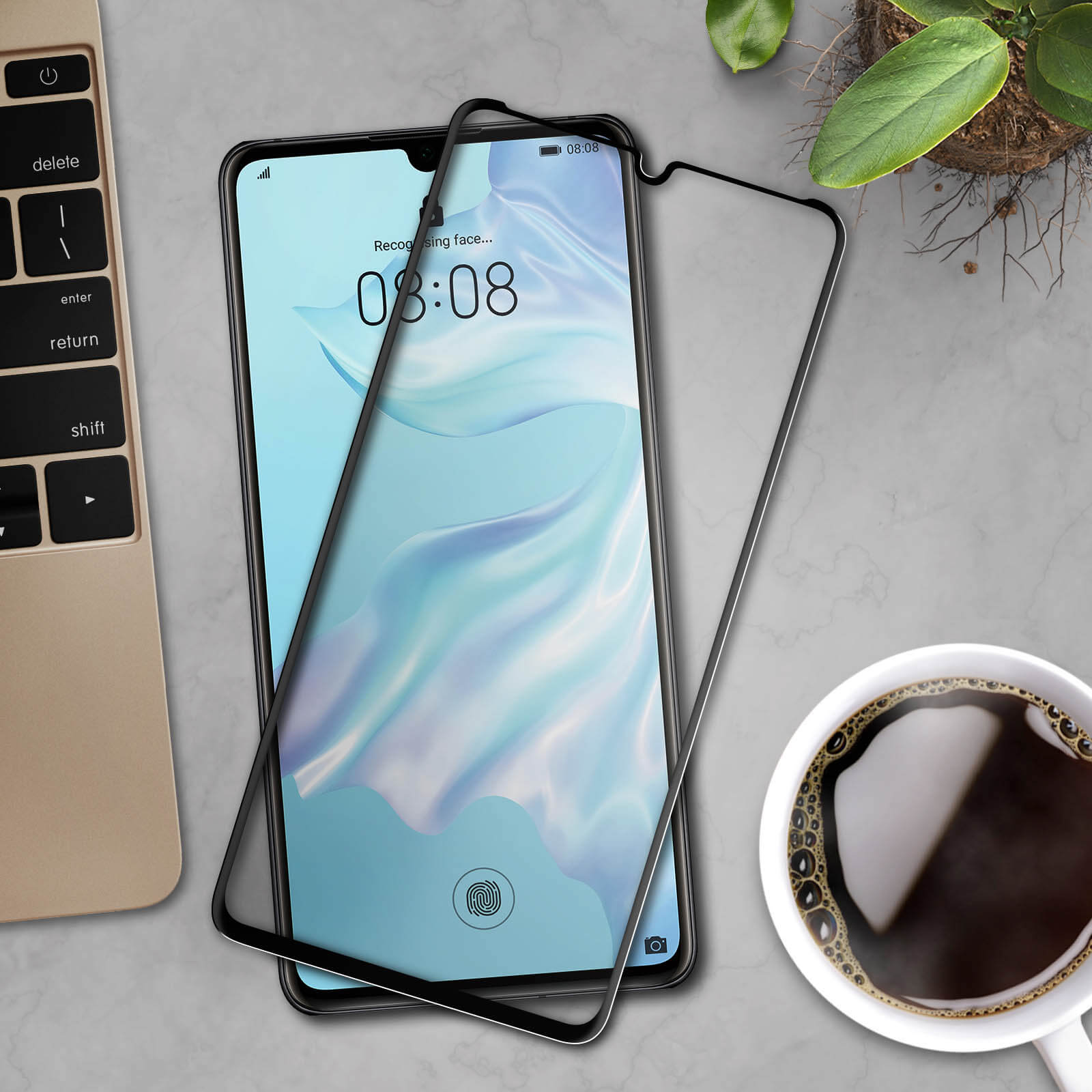 Uno smartphone con protezione schermo su un tavolo, con una tazza di caffè e una pianta, vista dall'alto.