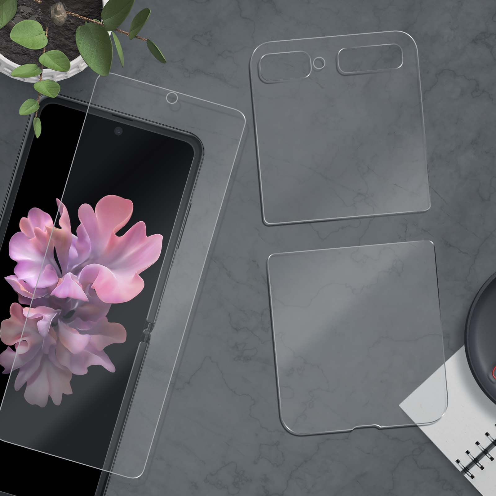 Telefono pieghevole con protezione schermo, piccola pianta e taccuino su una superficie grigia. Il telefono mostra un'immagine floreale rosa.