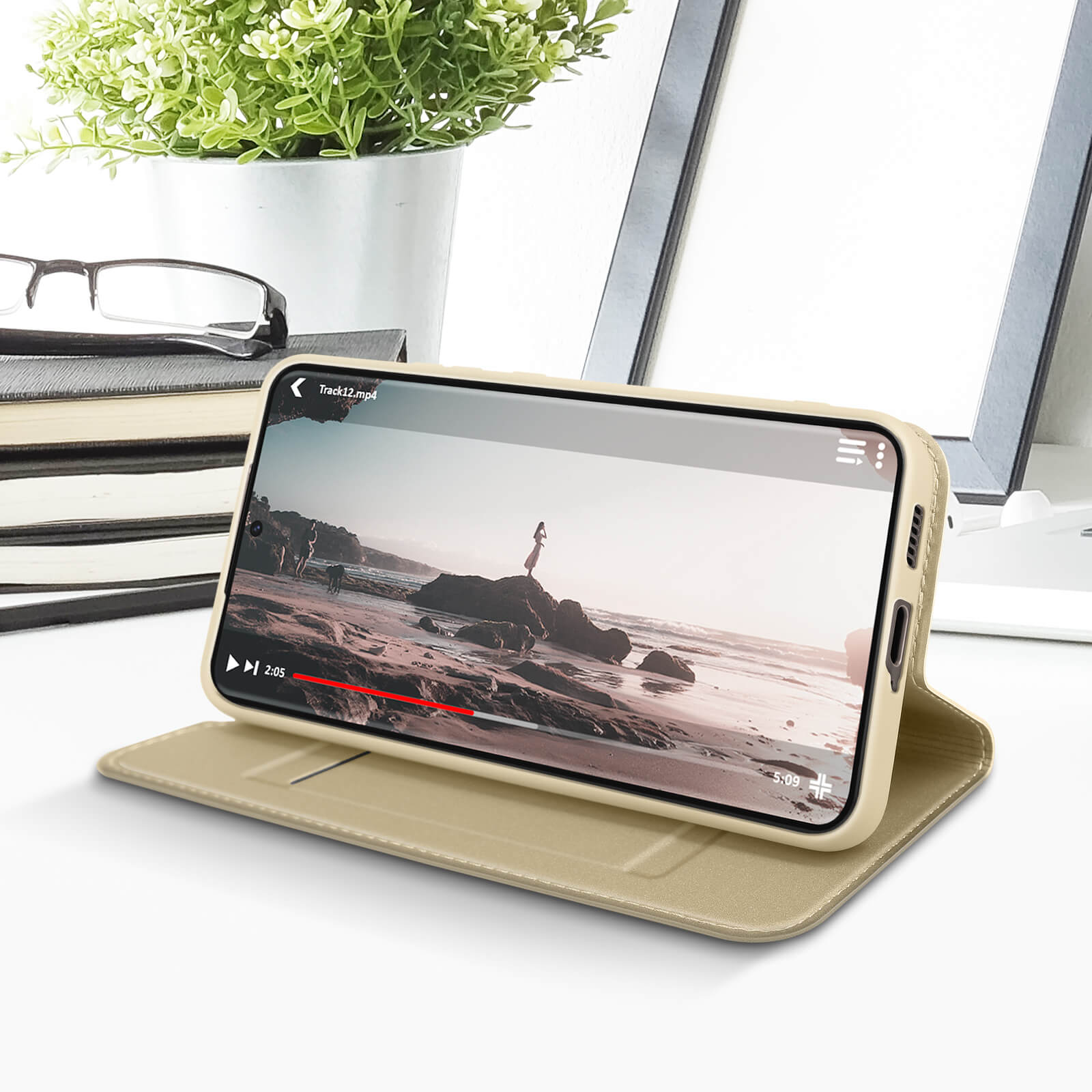 Telefono in custodia beige, che riproduce un video. Libri, occhiali e una pianta sullo sfondo.