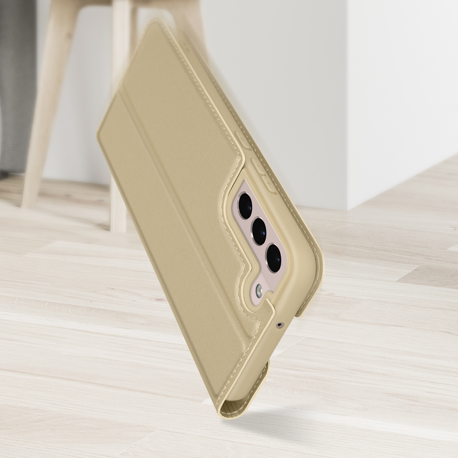 Telefono in custodia dorata, leggermente inclinato, contro pavimento in legno chiaro. Parete e sedia bianche sfocate.