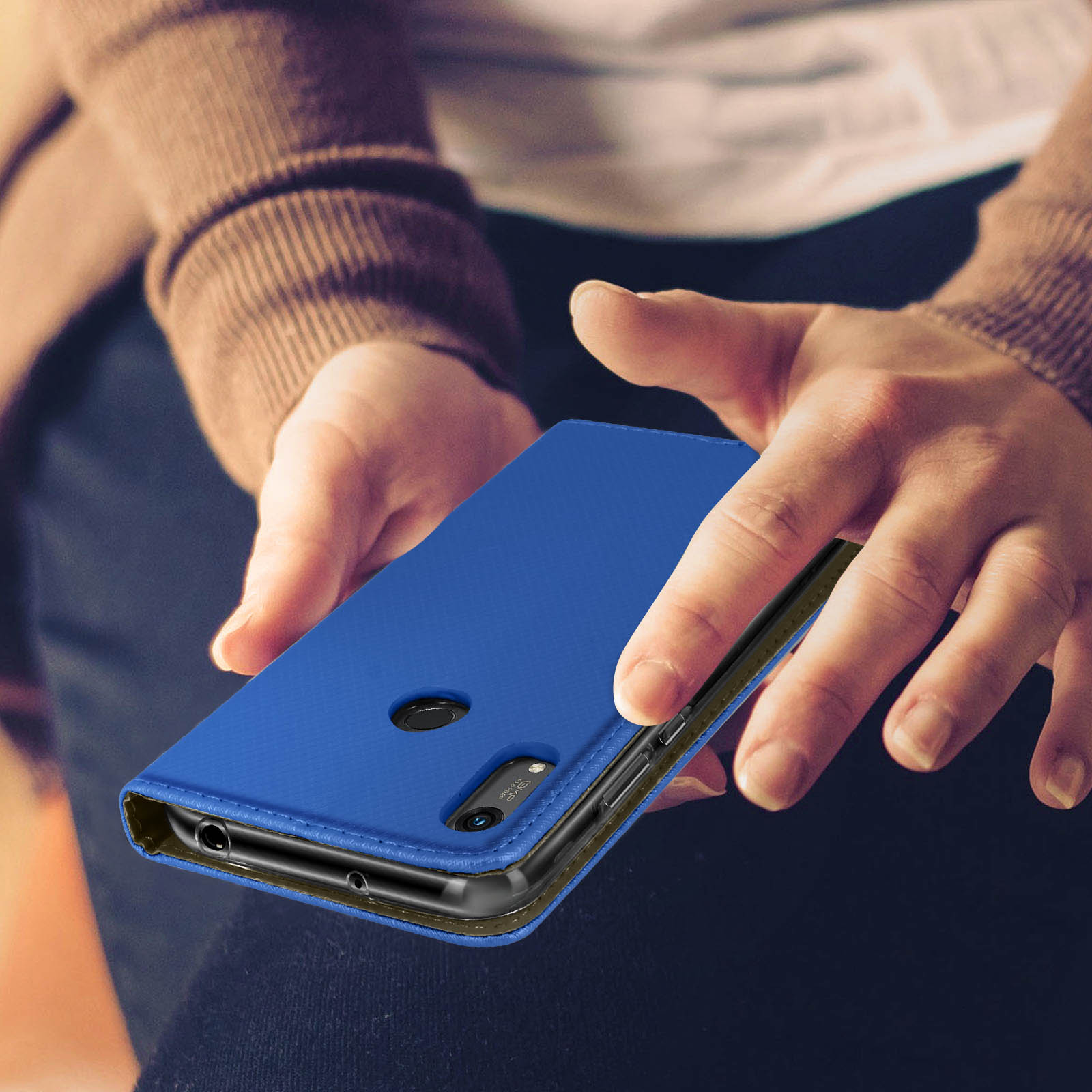 Un étui de téléphone bleu est tenu. Le téléphone est inséré. L'étui est texturé. La personne porte un pull marron.