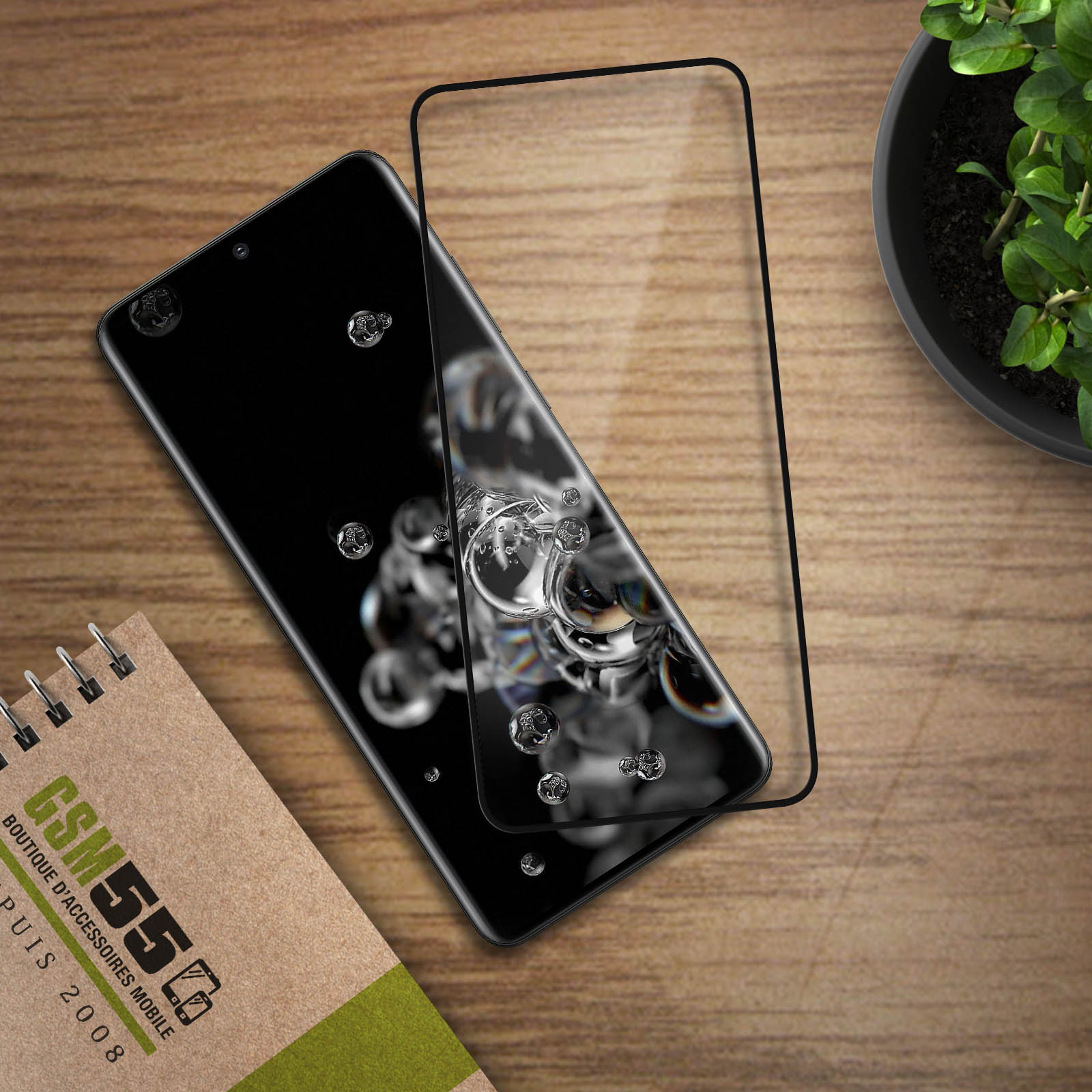 Smartphone nero con protezione schermo, gocce d'acqua, taccuino e pianta su tavolo di legno.