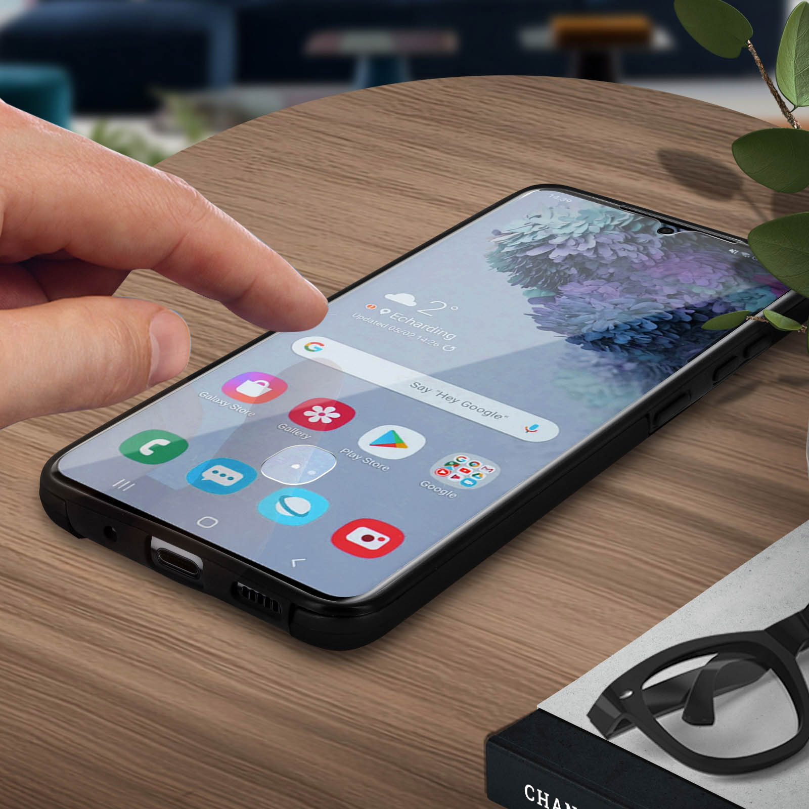 De vinger van een persoon raakt een telefoonscherm aan. Apps zijn zichtbaar. De telefoon ligt op een houten tafel.
