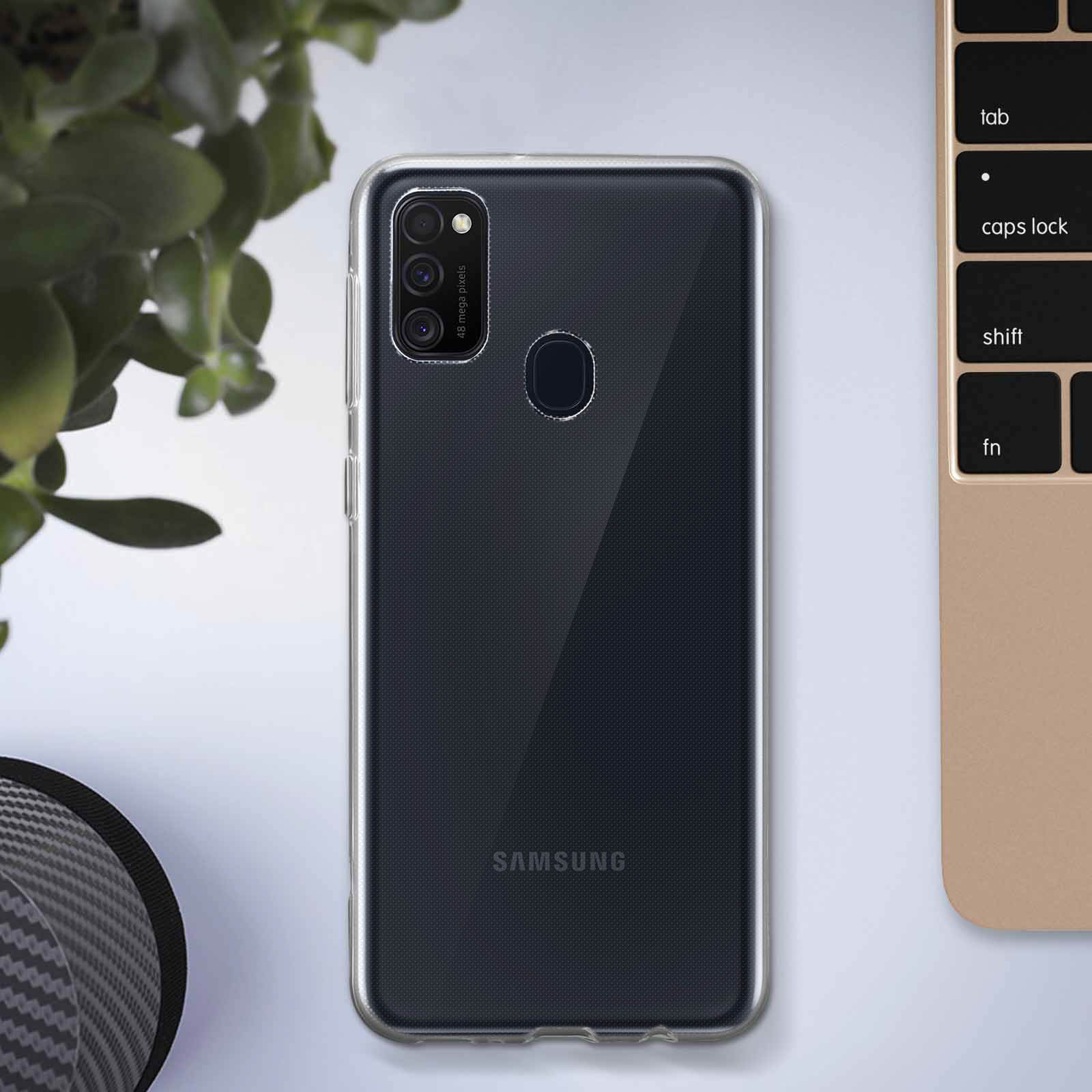 Telefono nero in custodia trasparente, logo Samsung visibile. Pianta sullo sfondo e tastiera a destra.