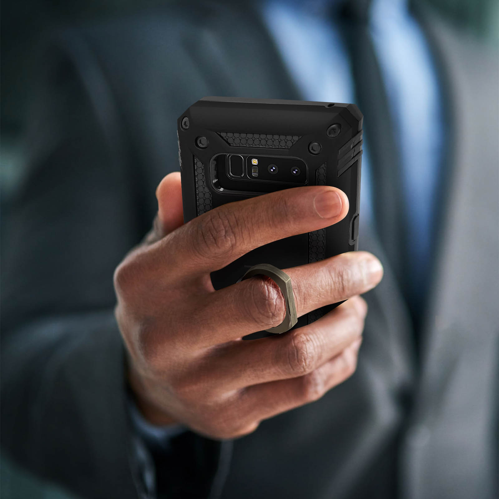 Person hält Telefon mit Ringhalter. Schwarze Hülle. Anzugjacke im Hintergrund.