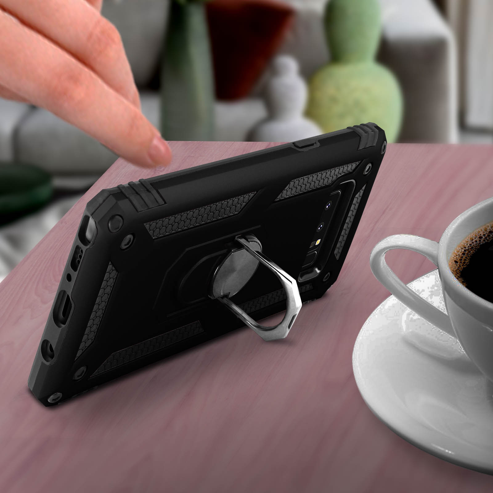 Schwarze Handyhülle mit Ringständer auf einem Tisch mit Kaffeetasse. Eine Hand greift nach dem Telefon.