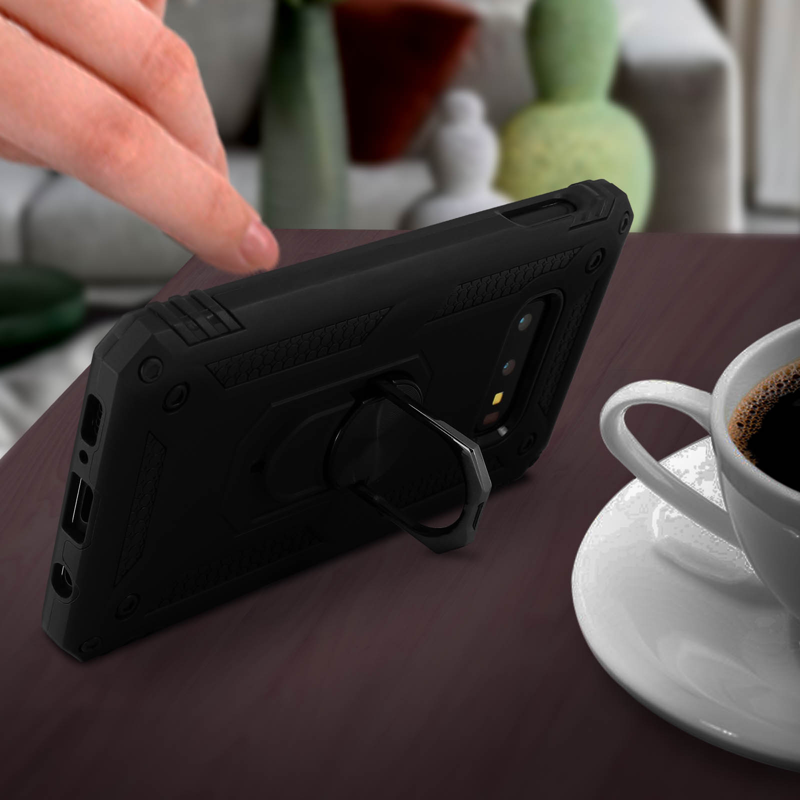 Coque de téléphone noire avec support de bague, sur table avec tasse à café et main. Arrière-plan flou avec objets.