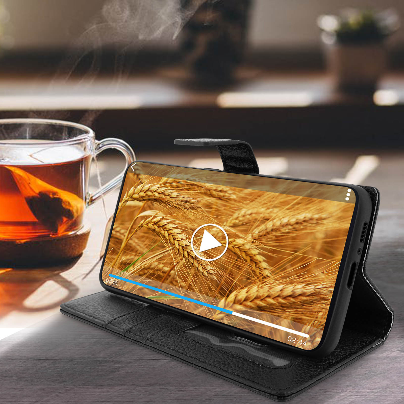 Telefono in custodia nera che mostra un video di grano, accanto a una tazza di tè e un tavolo di legno.