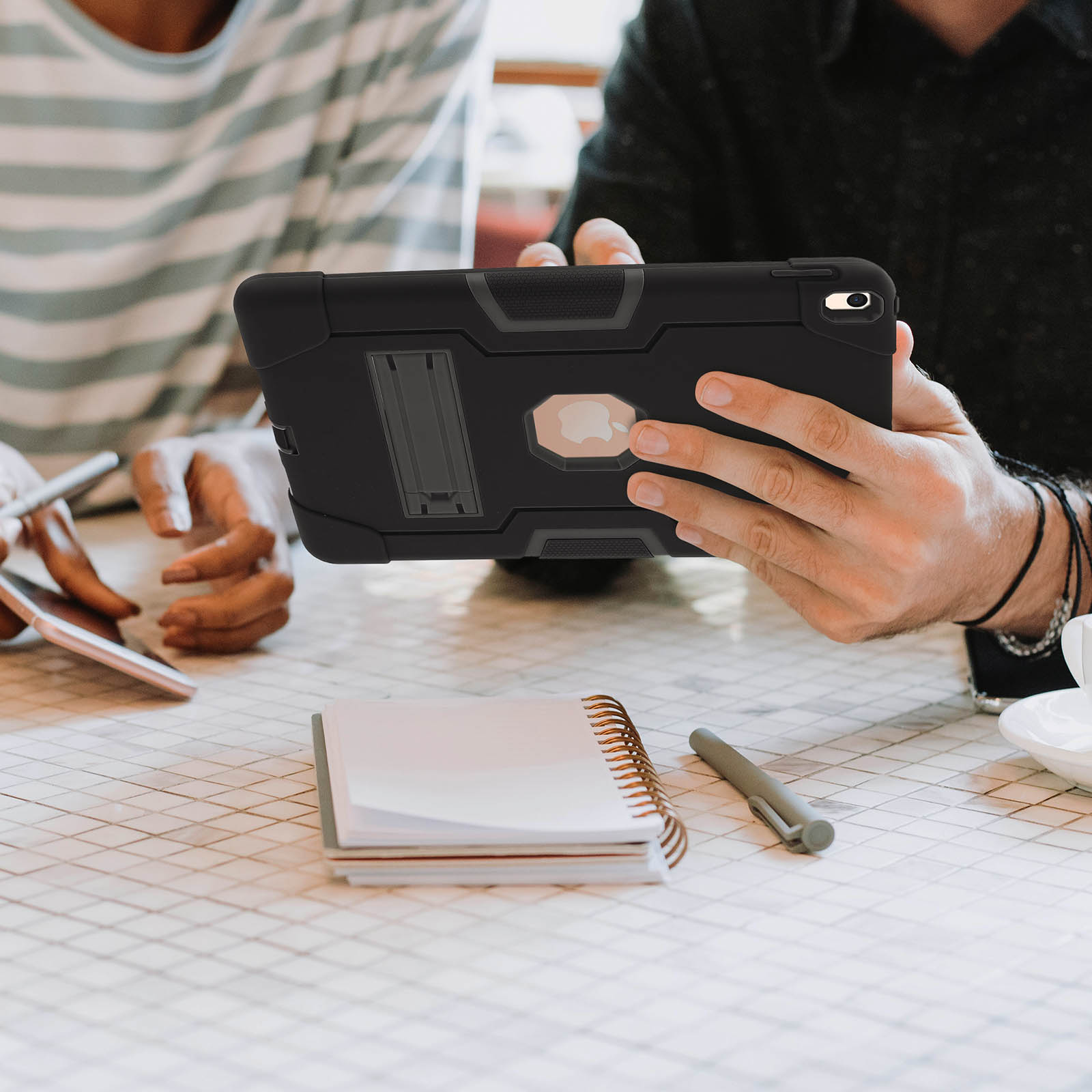 Personen an einem Tisch mit Tablet in schwarzer Hülle. Notizblock und Stift auf dem Tisch.