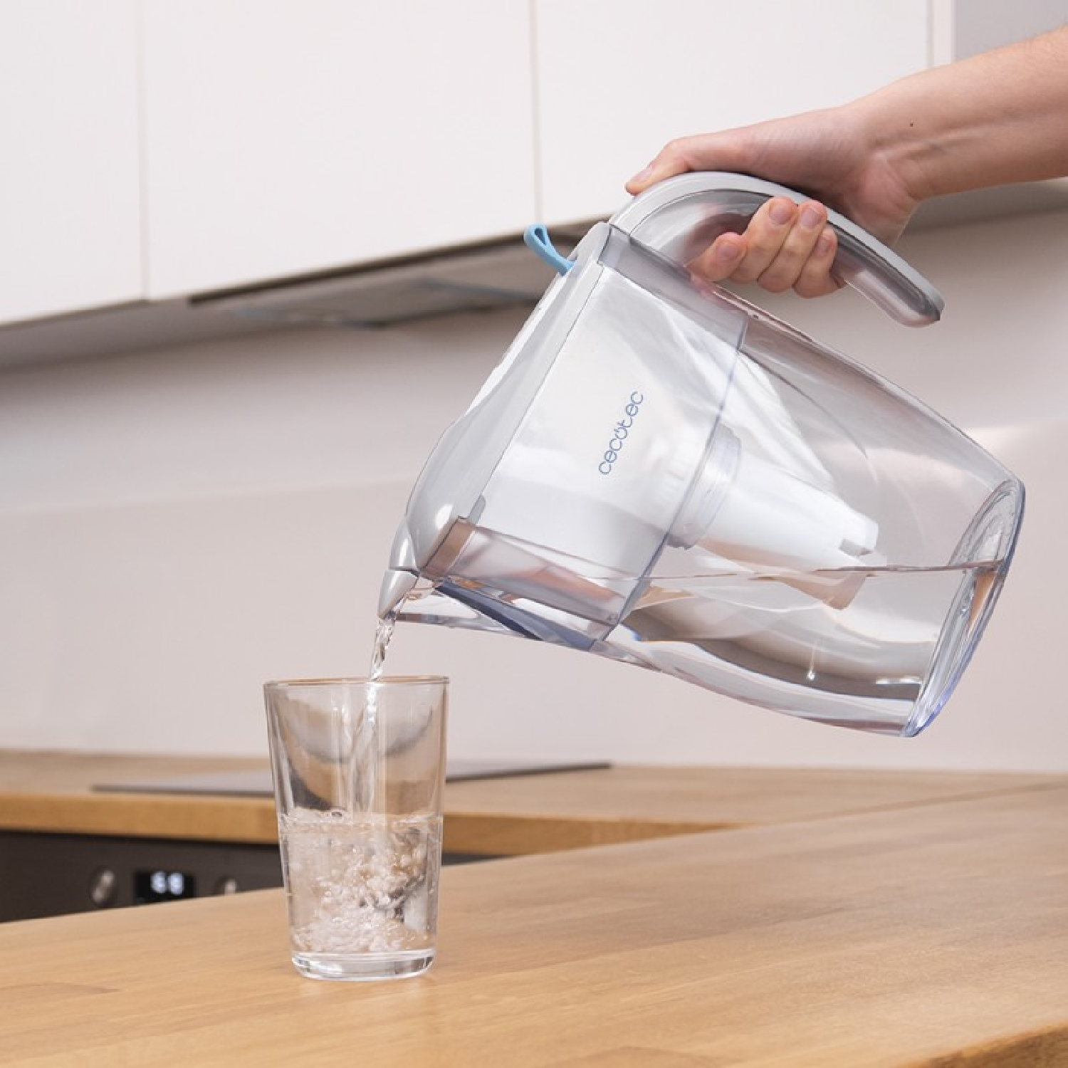 Una persona versa acqua da una caraffa filtrante in un bicchiere. Piano di lavoro in legno, cucina bianca.