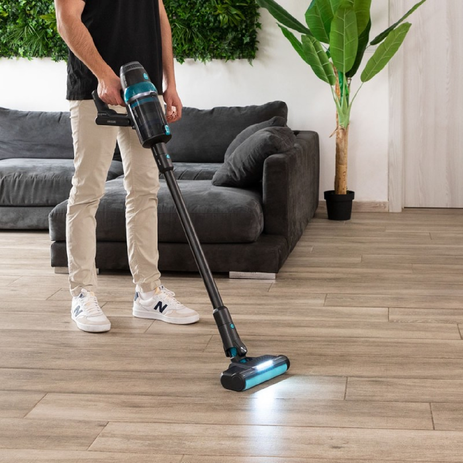 Un hombre aspira un piso de madera con una aspiradora inalámbrica negra y verde azulado. Un sofá y una planta en el fondo.