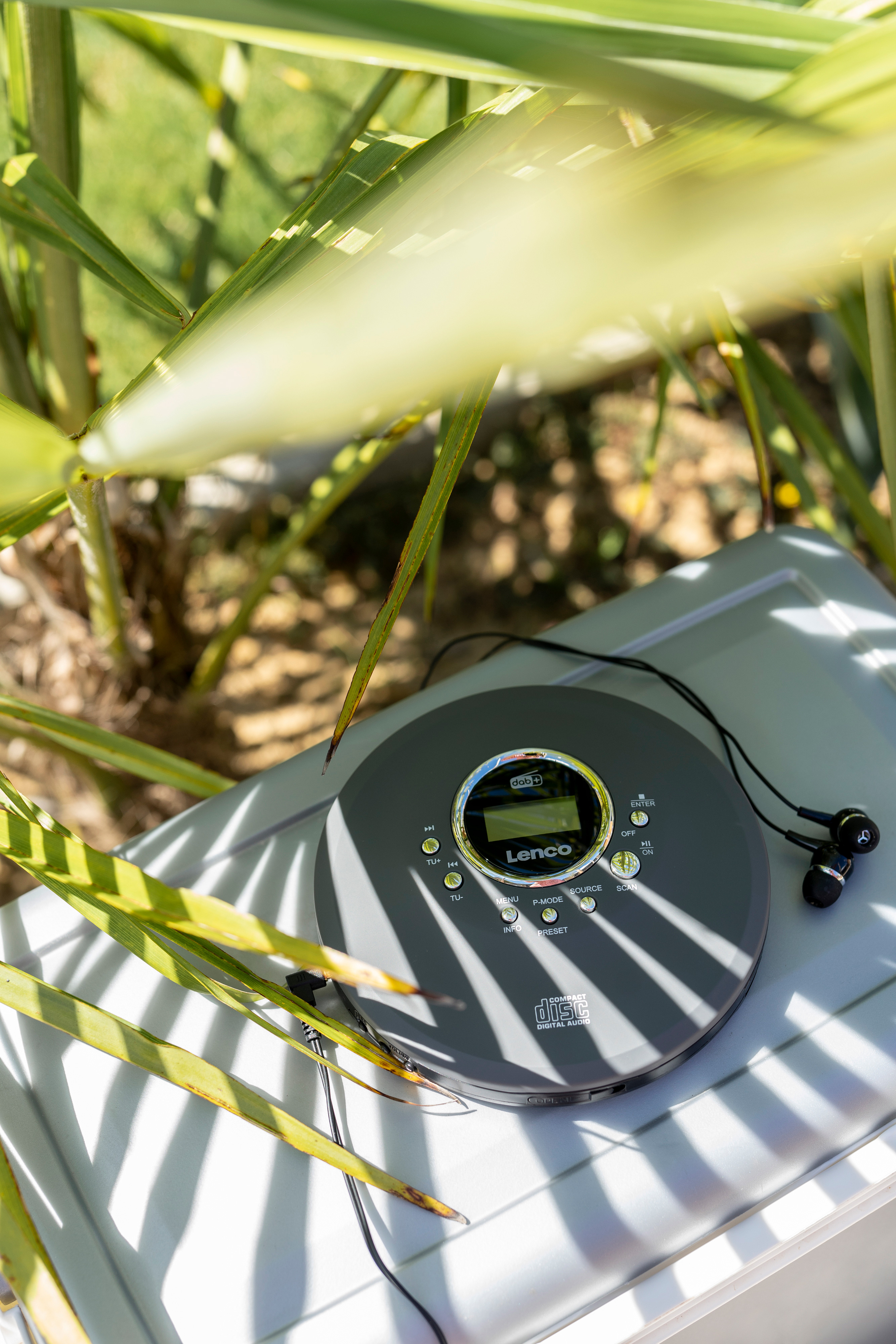Szary przenośny odtwarzacz CD ze słuchawkami, spoczywający na białej powierzchni wśród zielonych liści.