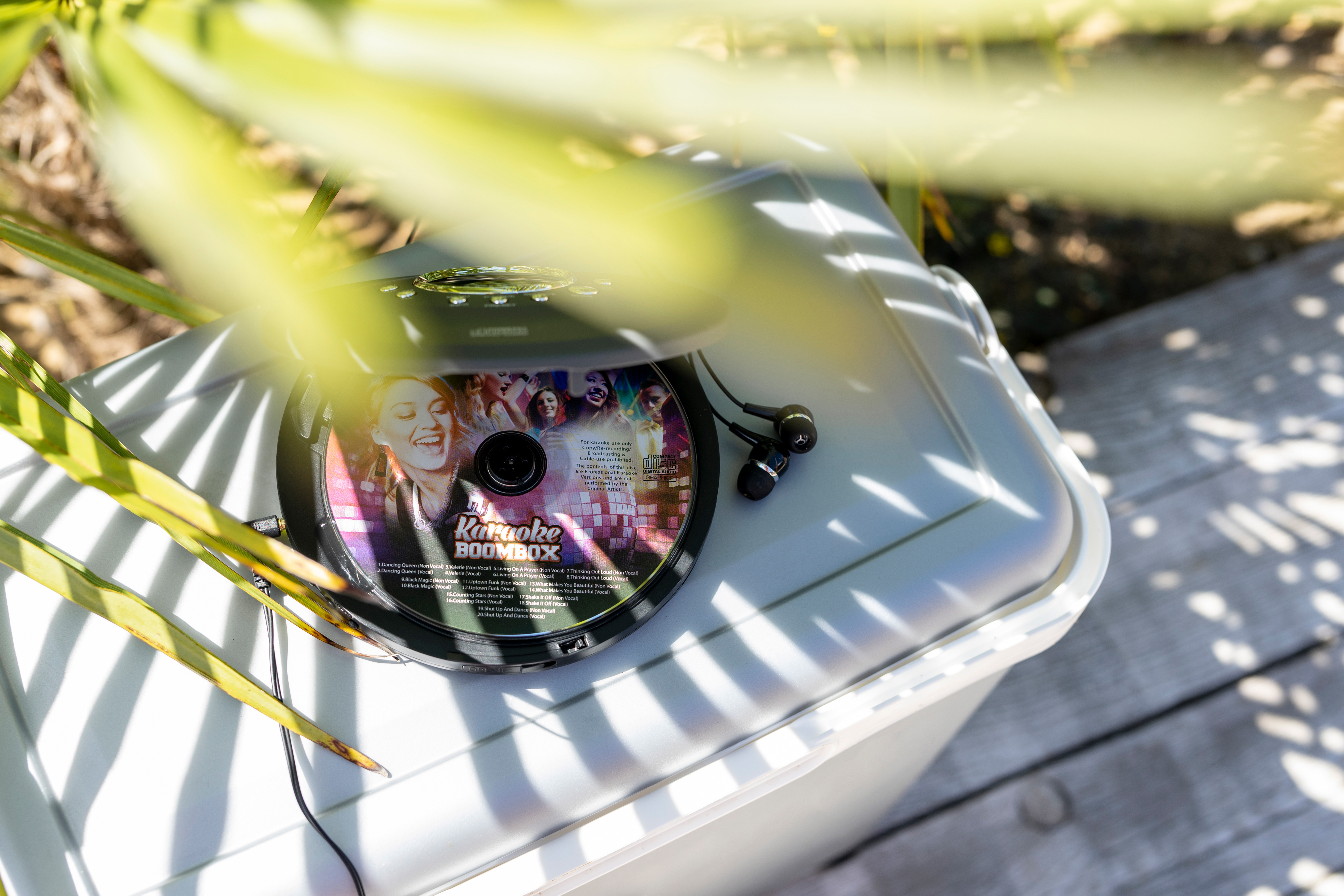 Odtwarzacz CD z płytą karaoke, słuchawkami i szarą skrzynką, z zielonymi liśćmi na pierwszym planie.