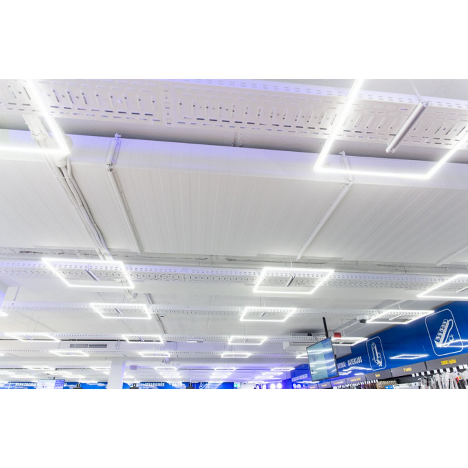 Ein Geschäftsraum mit weißer Decke und quadratischen LED-Leuchten, der Ladenzeilen und blaue Schilder in der Ferne zeigt.