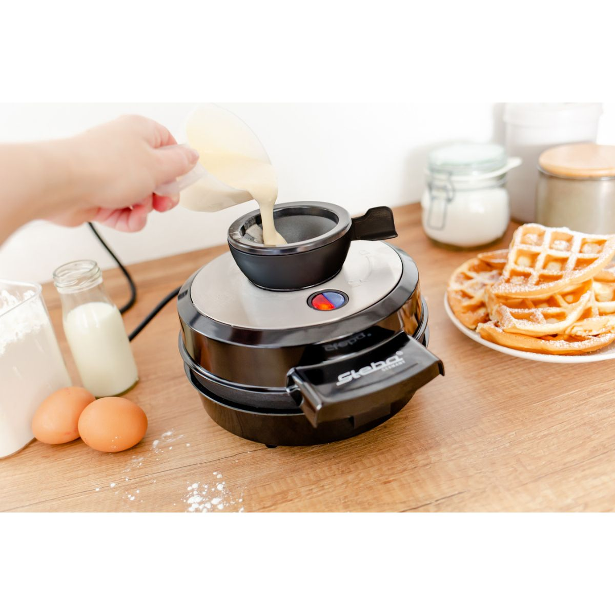 Eine Person gießt Teig in einen Waffeleisen, mit Waffeln in der Nähe.
