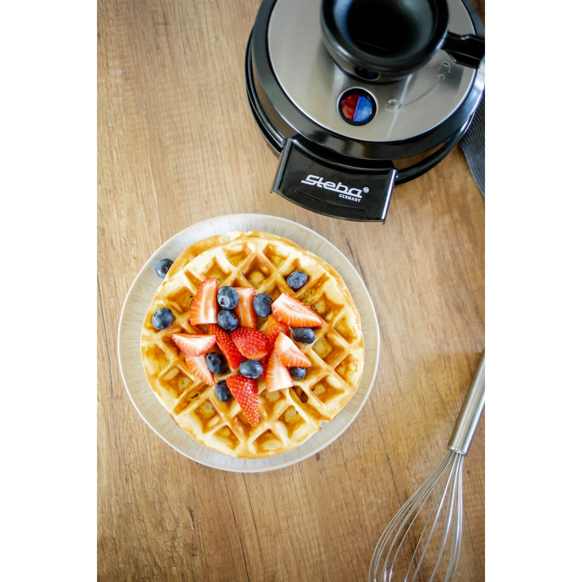 Waffeln mit Erdbeeren und Blaubeeren auf einem Teller, mit Waffeleisen und Schneebesen.