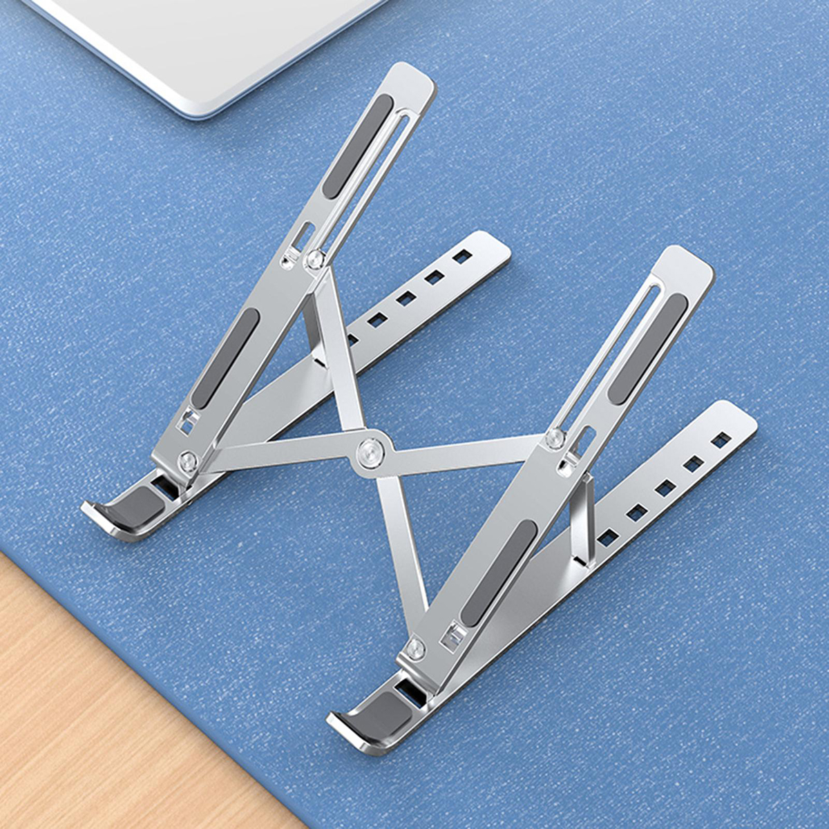 Soporte plateado para portátil sobre una superficie azul. El soporte está desplegado. Un portátil arriba a la izquierda.