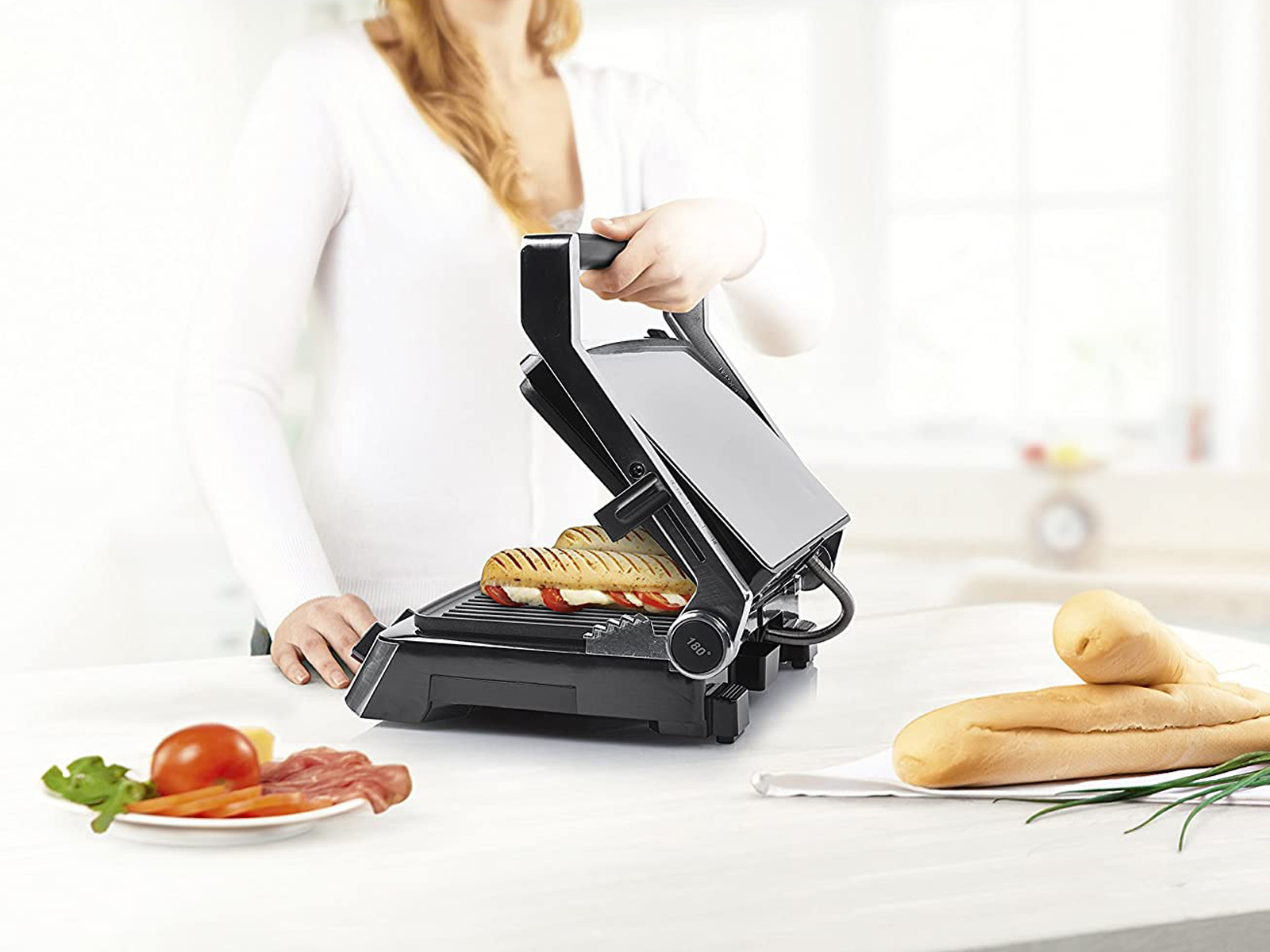 Eine Frau benutzt eine schwarze Panini-Presse. Ein Sandwich ist darin. Zutaten sind in der Nähe.