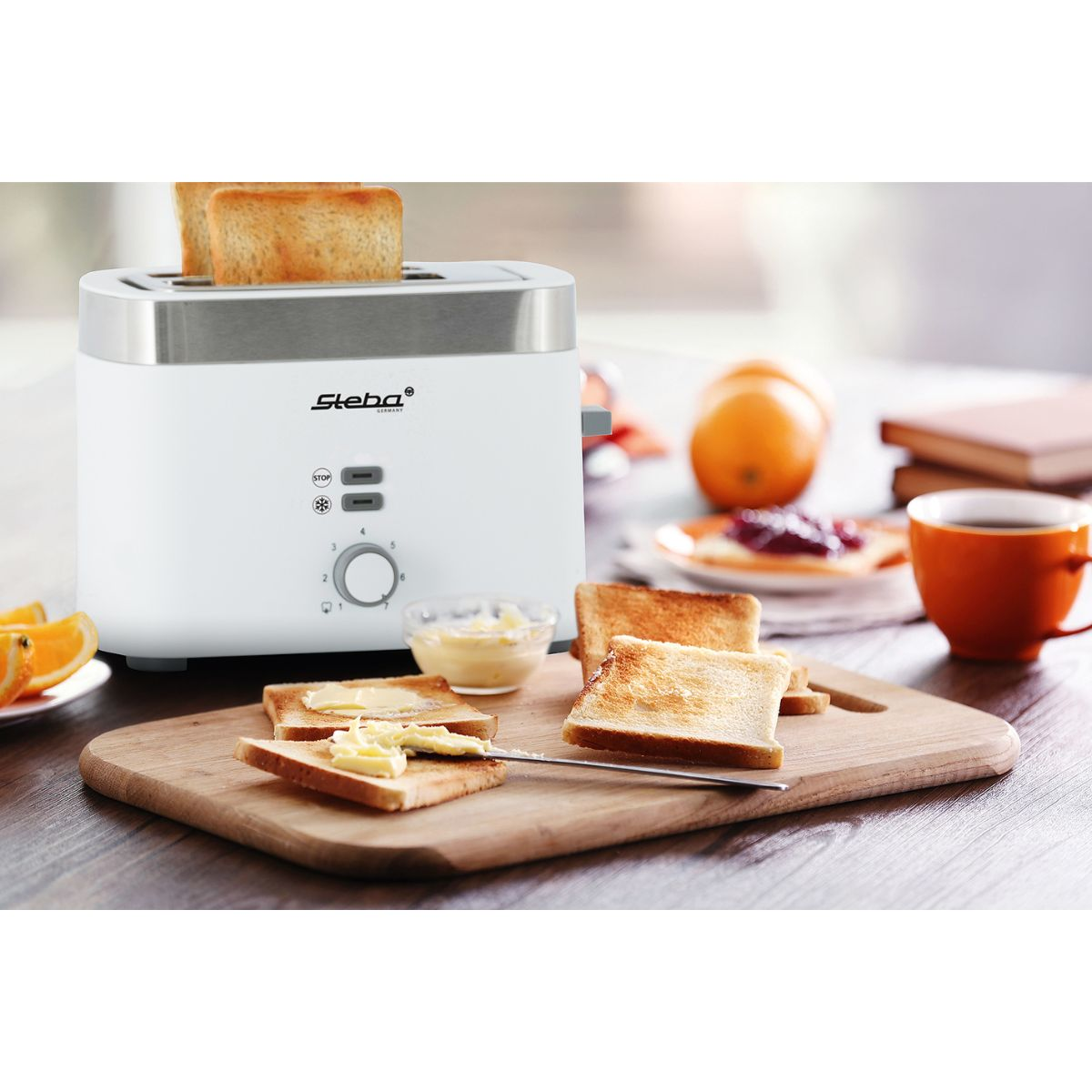 Weißer Toaster mit Toast, Butter und Orangen auf einem Holzbrett. Orangene Tasse daneben.