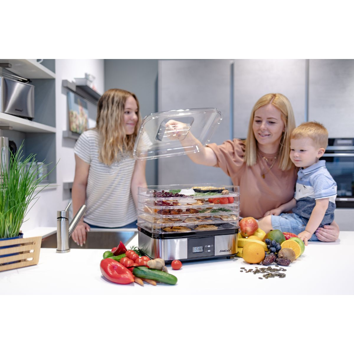 Ein Lebensmittel-Dehydrator auf einer Küchentheke. Zwei Frauen und ein Kind sind anwesend.