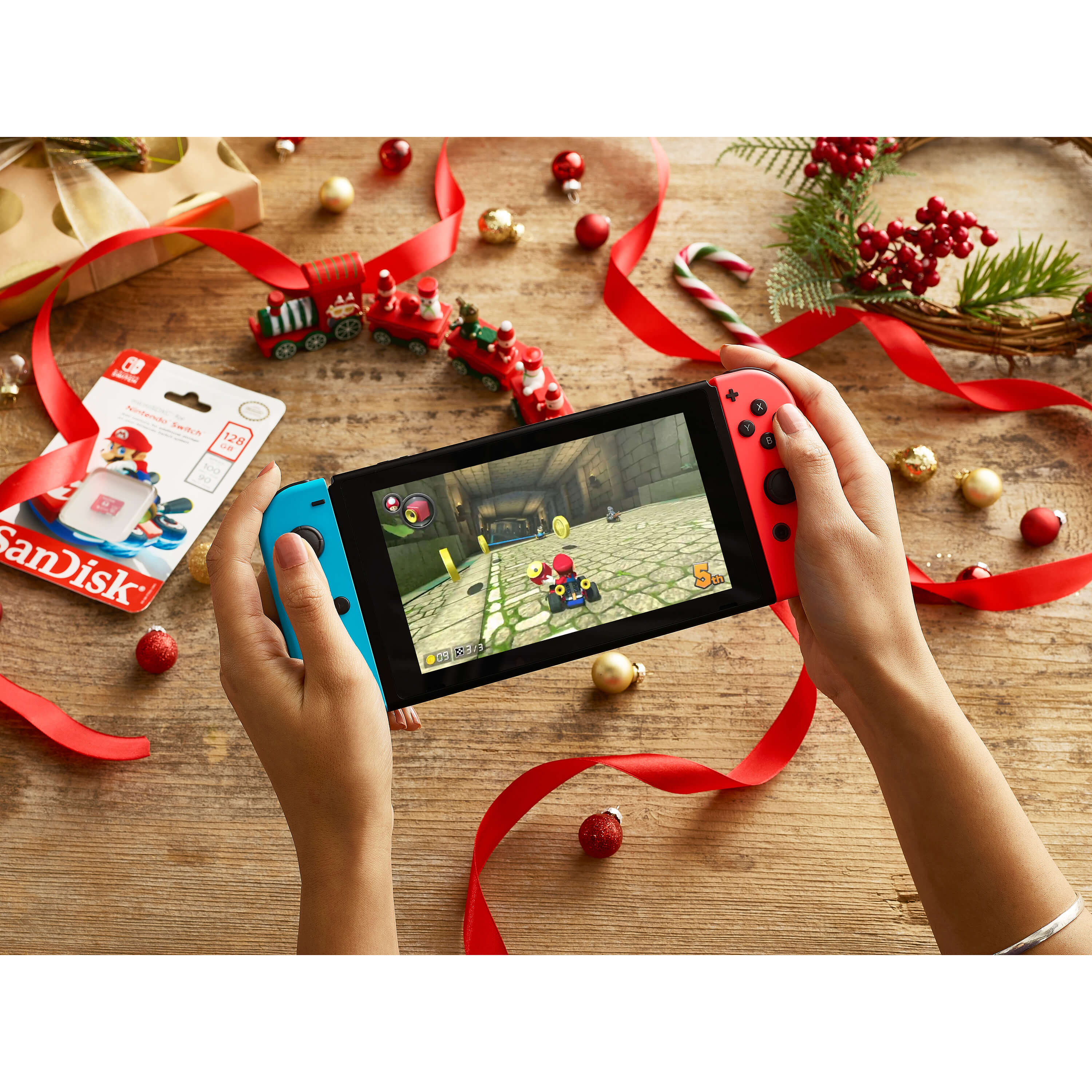 Mani che tengono una Nintendo Switch con un gioco sullo schermo, circondata da decorazioni natalizie e una confezione regalo.