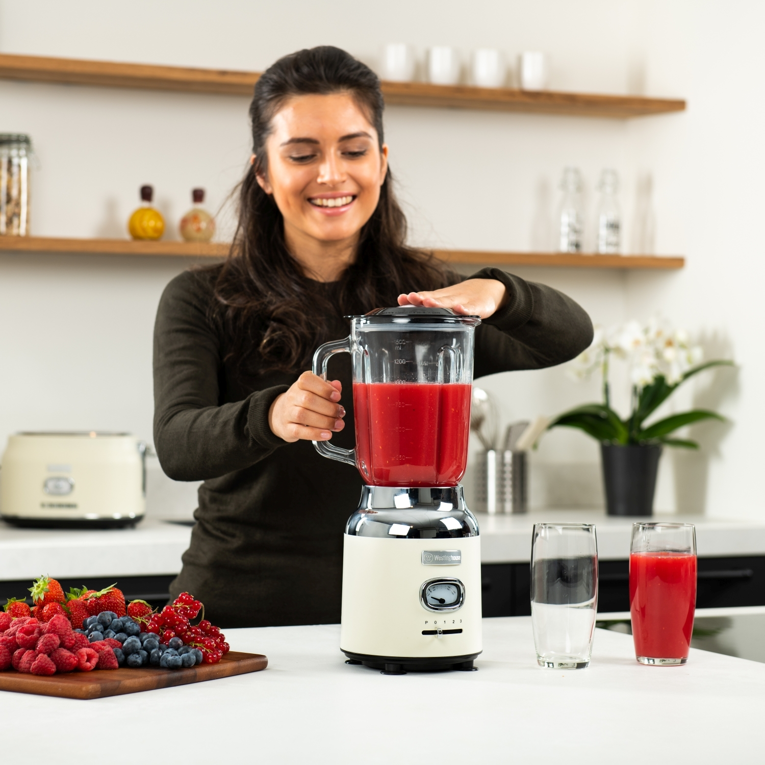 Frau, die einen roten Smoothie in einem weißen und cremefarbenen Mixer mixt, mit Früchten auf dem Tisch.