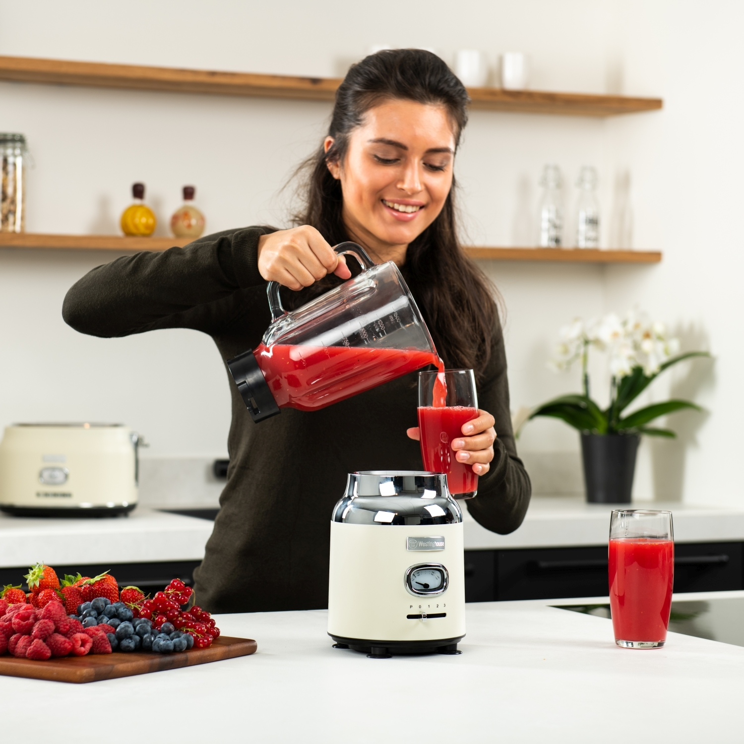 Eine Frau gießt rote Flüssigkeit aus einem Mixer in ein Glas, neben einem Mixer und Früchten auf einem weißen Tisch.