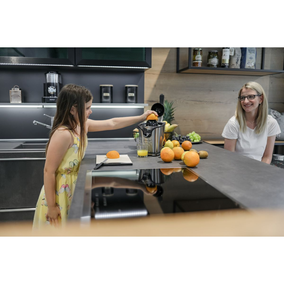 Una niña pone naranja en exprimidor, mujer mirando en una cocina moderna.